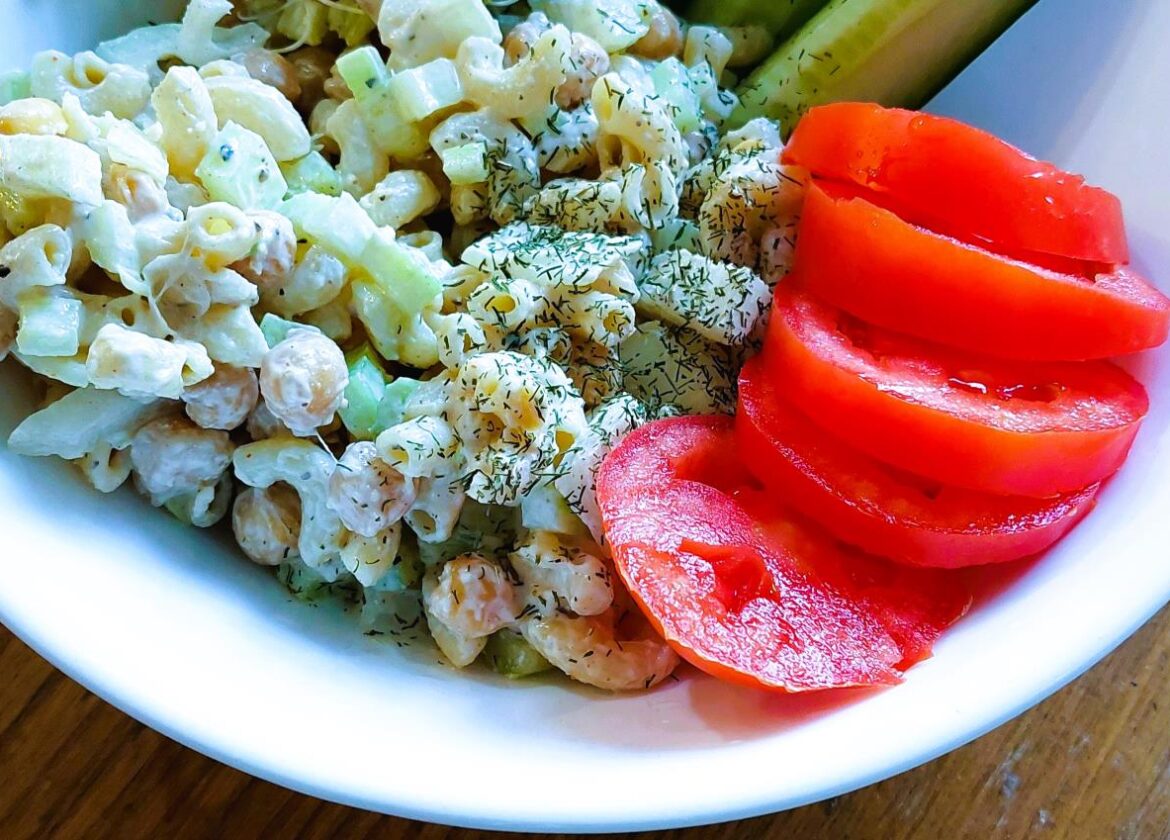 Chickpea 'Tuna' Salad served in a white bowl alongside sliced red tomatoes, cucumber wedges, and fresh broccoli sprouts on a wooden surface.