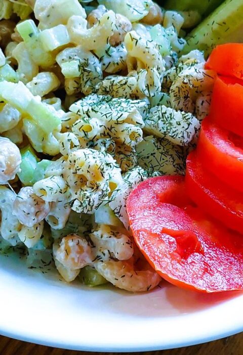 Chickpea 'Tuna' Salad served in a white bowl alongside sliced red tomatoes, cucumber wedges, and fresh broccoli sprouts on a wooden surface.