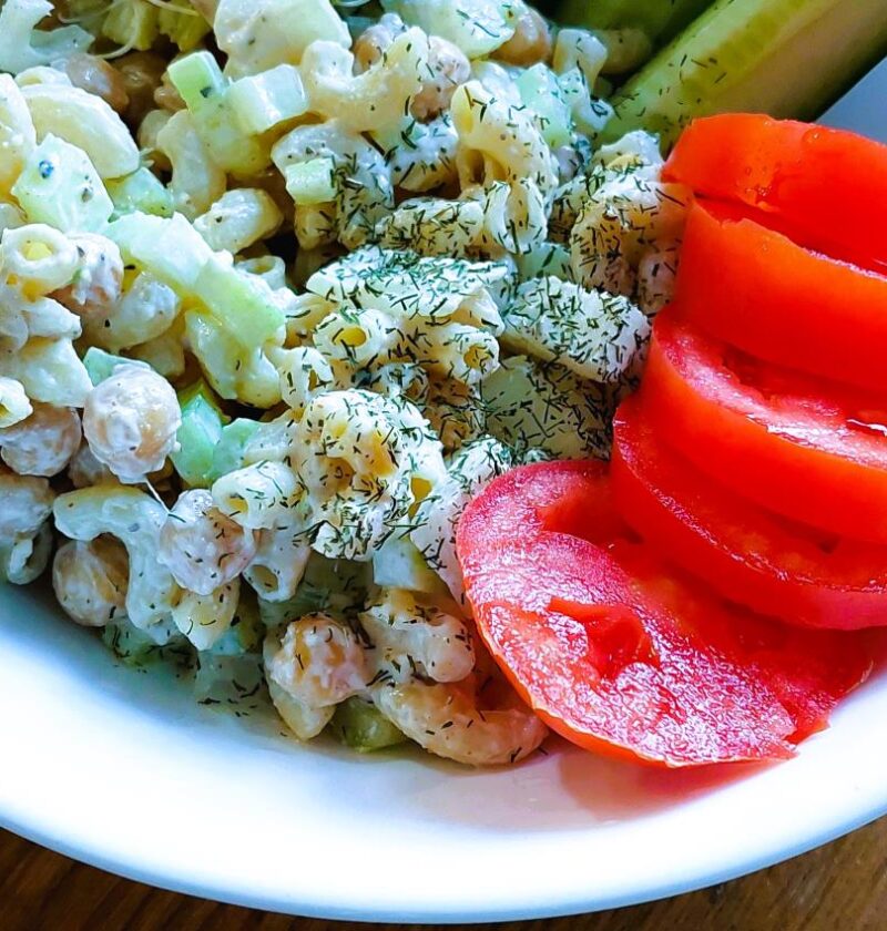Chickpea 'Tuna' Salad served in a white bowl alongside sliced red tomatoes, cucumber wedges, and fresh broccoli sprouts on a wooden surface.