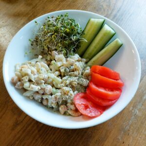 Chickpea 'Tuna' Salad served in a white bowl alongside sliced red tomatoes, cucumber wedges, and fresh broccoli sprouts on a wooden surface.
