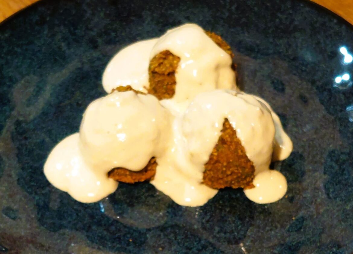 Three falafels covered in creamy tahini sauce served on a handmade dark blue glazed plate.