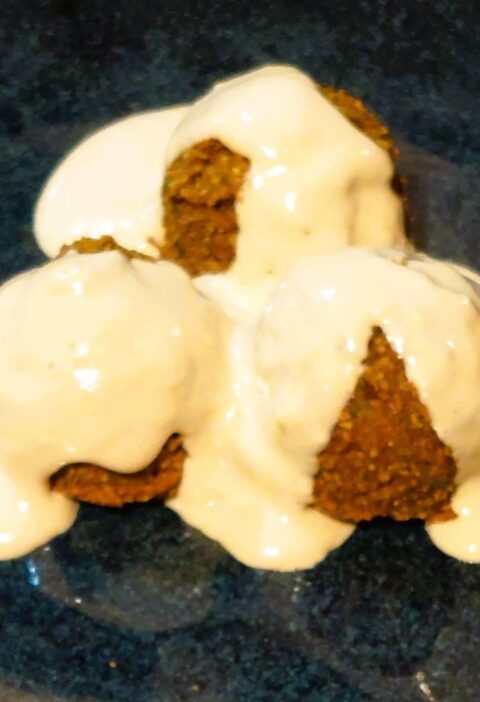 Three falafels covered in creamy tahini sauce served on a handmade dark blue glazed plate.