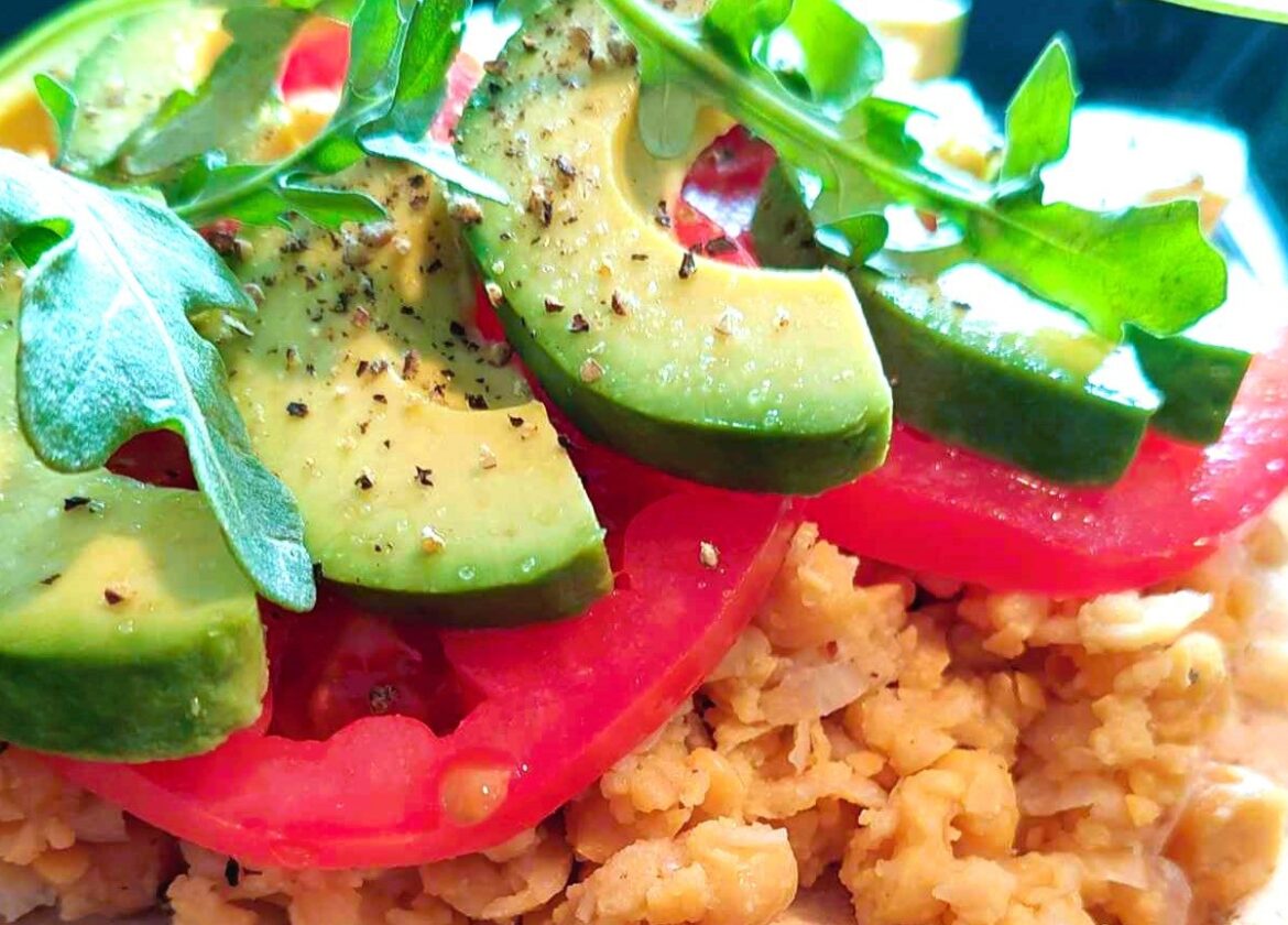 Close-up of a protein-packed plant-based meal featuring mashed chickpeas topped with sliced avocado, fresh tomato, and arugula, seasoned with salt and black pepper.