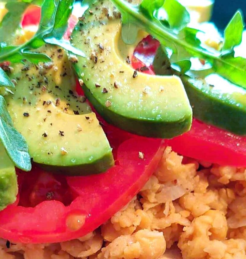 Close-up of a protein-packed plant-based meal featuring mashed chickpeas topped with sliced avocado, fresh tomato, and arugula, seasoned with salt and black pepper.